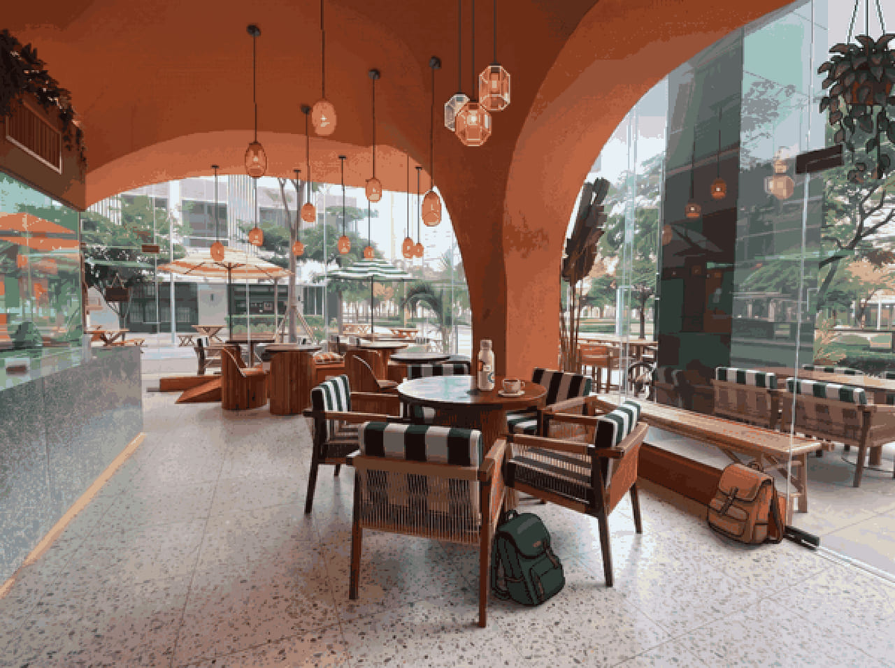 A cafe terrace under terracotta arches, glass walls onto a tree-lined street; a water bottle and a coffee cup on a round table, one backpack under a chair and another by the bench.